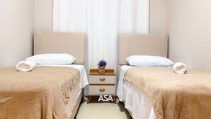 A hotel room with two beds, each with a white pillow and a beige blanket. There is a small wooden nightstand with a drawer between the beds. - ASA Verona Park 06 2 Bedrooms Single Story House Timbo (Timbo)