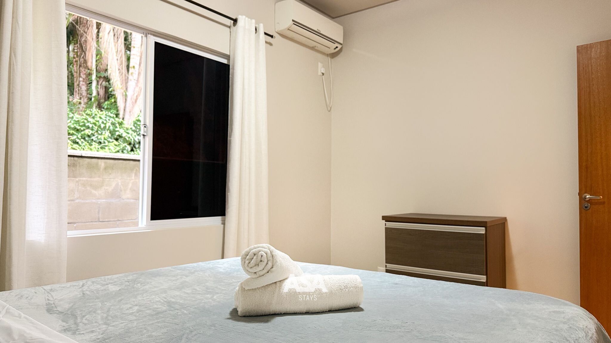 Una habitación de hotel con cama, mesita de noche, aire acondicionado y ventana con cortinas.