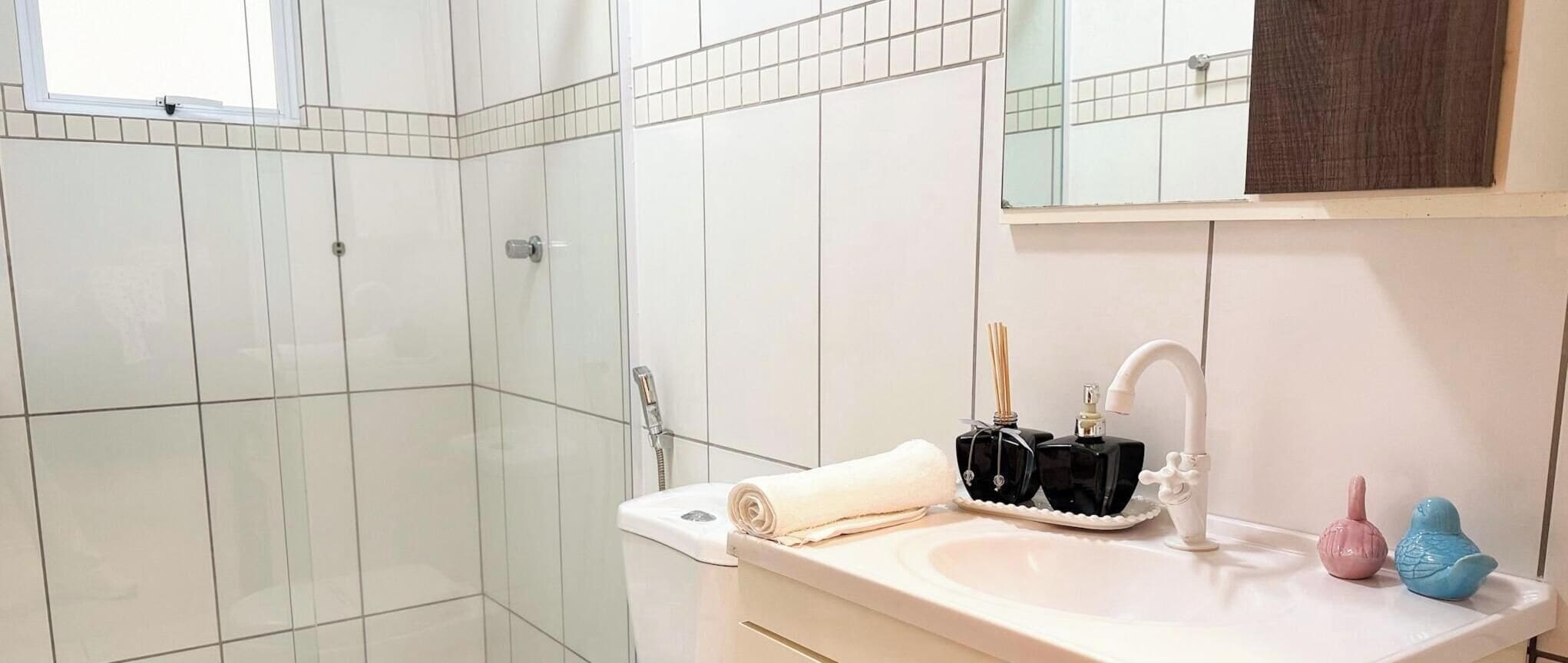 Casa de banho com azulejos brancos, pia com torneira, toalha e dois dispensadores de sabonete em formato de pássaros.