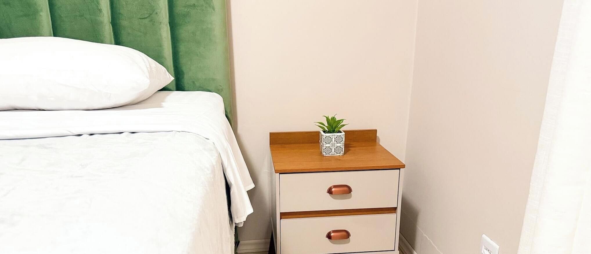 A bed with white bedding, a green headboard, and a nightstand with drawers and a small plant.