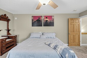 A bedroom with a bed, a dresser, a ceiling fan, a door, and two wall-mounted paintings. - GREENVILLE GETAWAY:  (Greenville, MI) Lakefront on Perch Lake, Sleeps 6-Kayak, Paddle Boat-Dock! (Greenville)