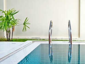 Ein Schwimmbecken mit zwei Metallhandläufen, einer kleinen Pflanze und einer weißen Wand im Hintergrund.