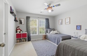 Ein Schlafzimmer mit einem Bett, einem Nachttisch, einem Bücherregal, einem Fenster mit Vorhängen und einem Deckenventilator.