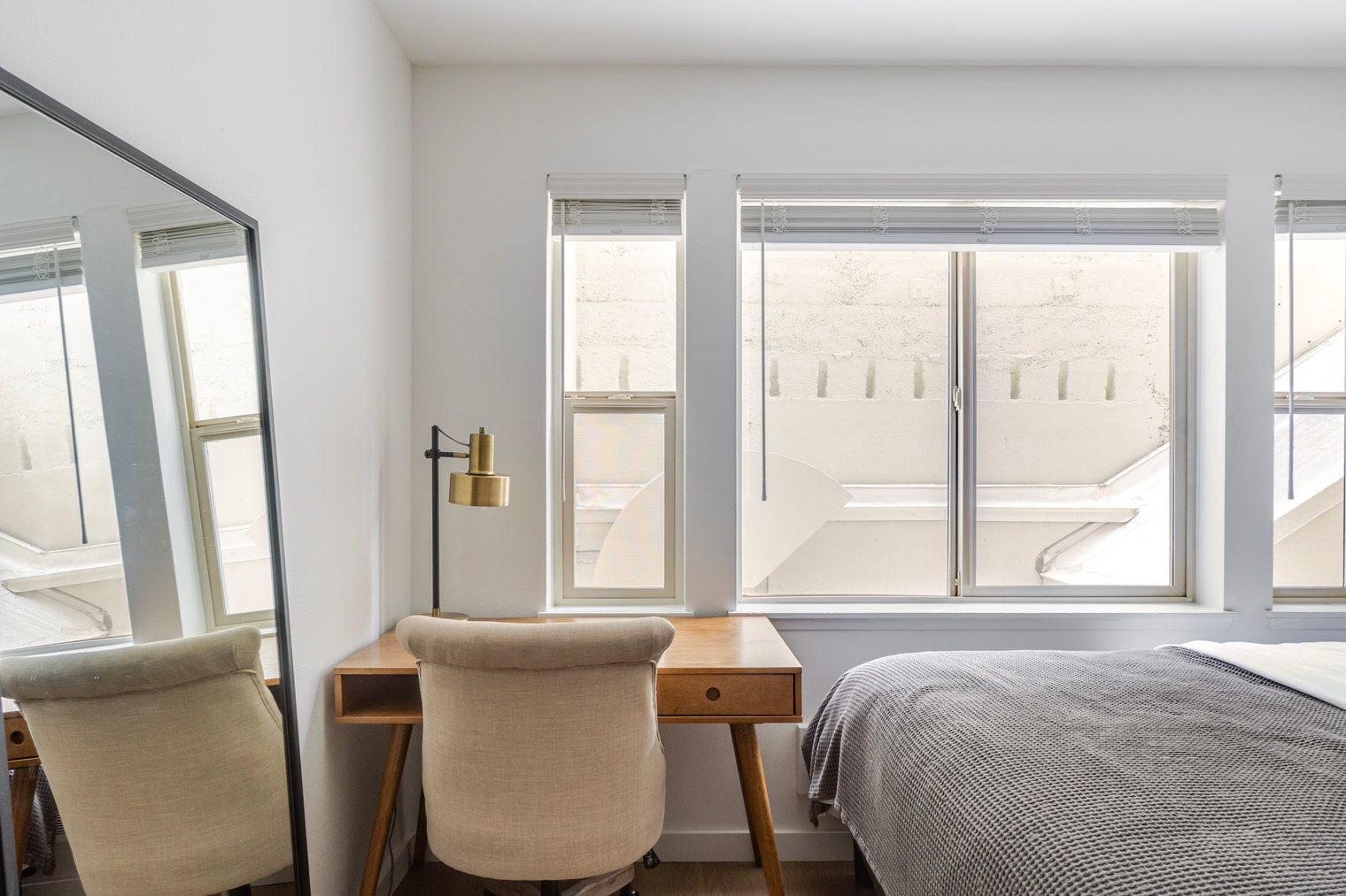 Quarto com cama, escrivaninha, cadeira e uma grande janela com vista para um edifício.