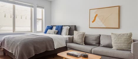 A modern bedroom with a bed, a grey sofa, a wooden coffee table, and a large window.