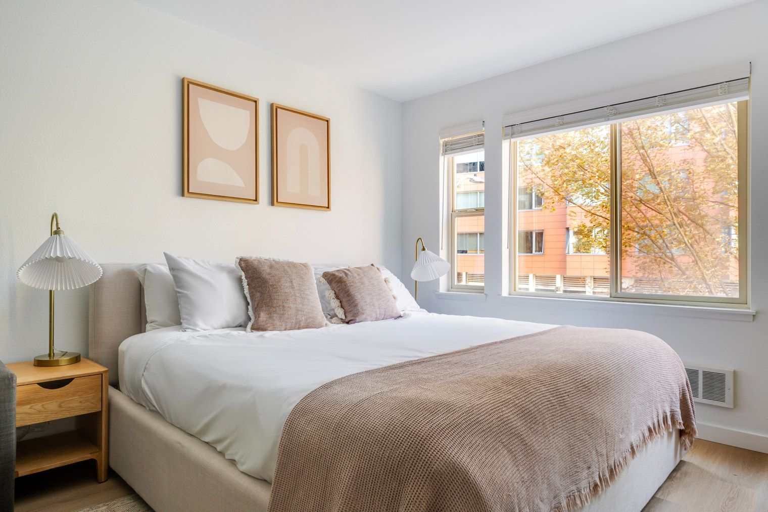 A bedroom with a large bed, two bedside lamps, a window with a view of buildings, and a wall with two framed pictures.