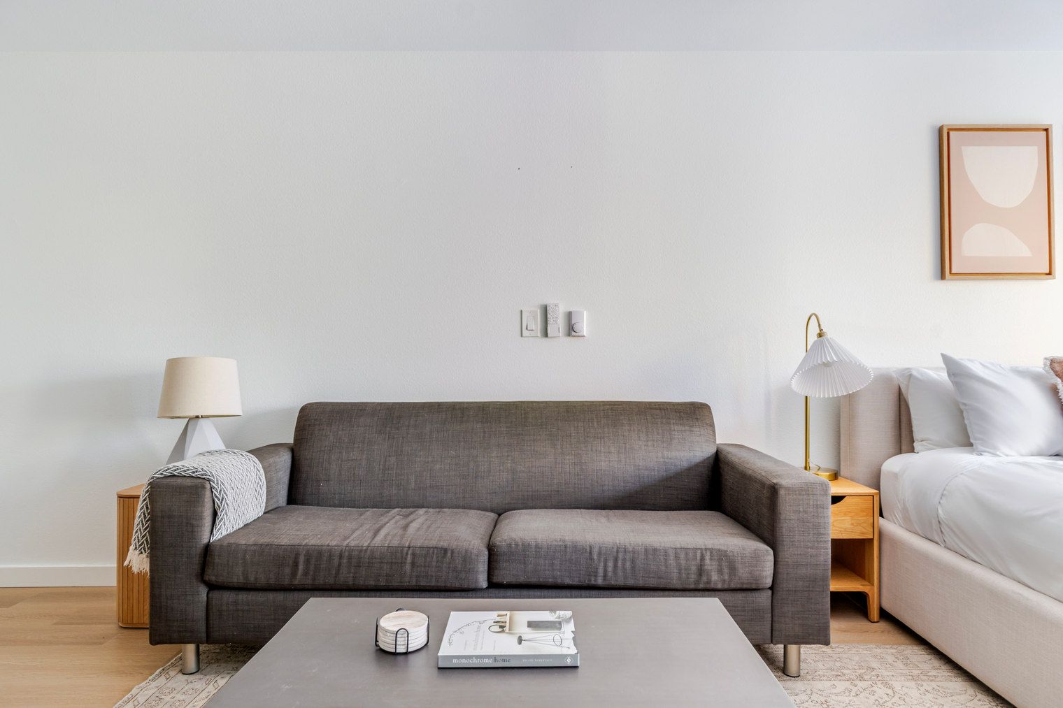 Ein modernes Wohnzimmer mit einem grauen Sofa, einem Couchtisch mit einem Buch, einer Lampe und einem Bett mit weißer Bettwäsche.