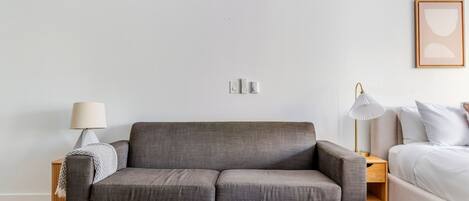 A modern living room with a grey sofa, a coffee table with a book, a lamp, and a bed with white bedding.
