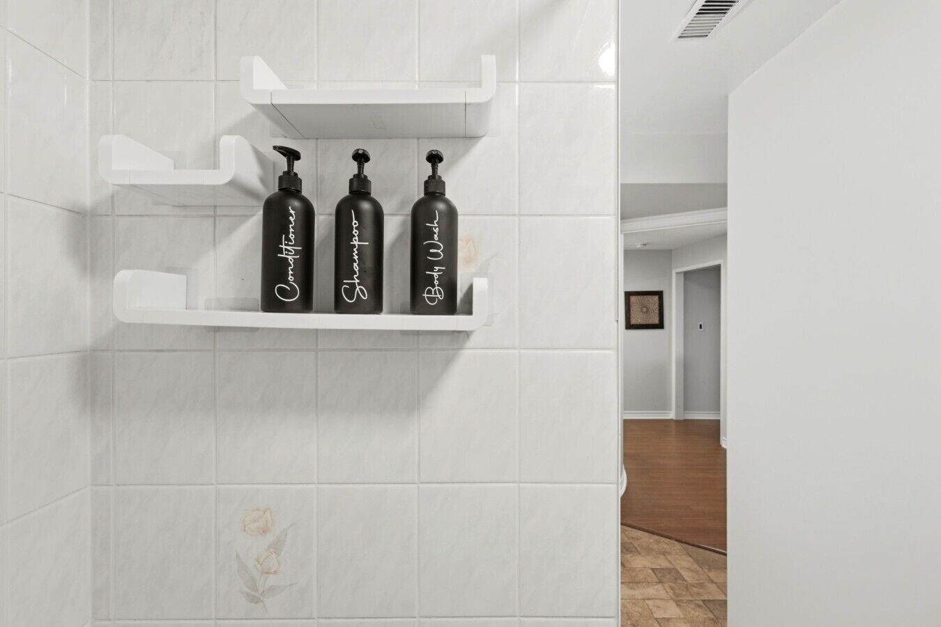 Une salle de bain avec des murs recouverts de carreaux blancs et une étagère sur laquelle reposent trois bouteilles noires avec des inscriptions blanches.