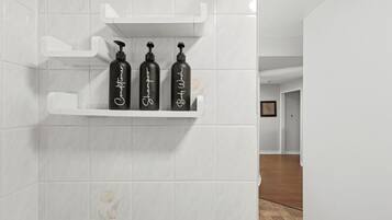 A bathroom with white tiled walls and a shelf holding three black bottles with white text.