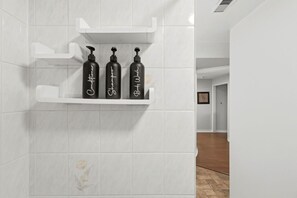 Casa de banho com paredes em azulejos brancos e uma prateleira com três frascos pretos e texto branco.