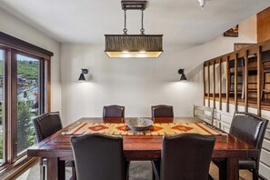 Une salle à manger avec une grande table en bois, des chaises en cuir, un tapis à motifs et des luminaires suspendus.