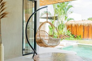 Una silla colgante junto a un ventanal de vidrio, una mesa redonda y una piscina con una estructura de techo de paja al fondo.