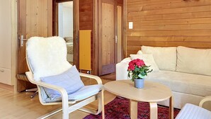 Een kamer met een houten vloer, een witte stoel met een zacht kussen, een beige bank, een kleine ronde tafel met een bloevas, en houten deuren.