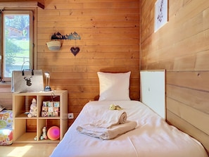 Habitación con paneles de madera, una cama, una mesita de noche, un estante con juguetes y una ventana con vista a árboles.