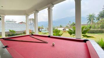 A pool table with a cue stick and a billiard ball, set against a backdrop of a garden with palm trees and a mountainous landscape.