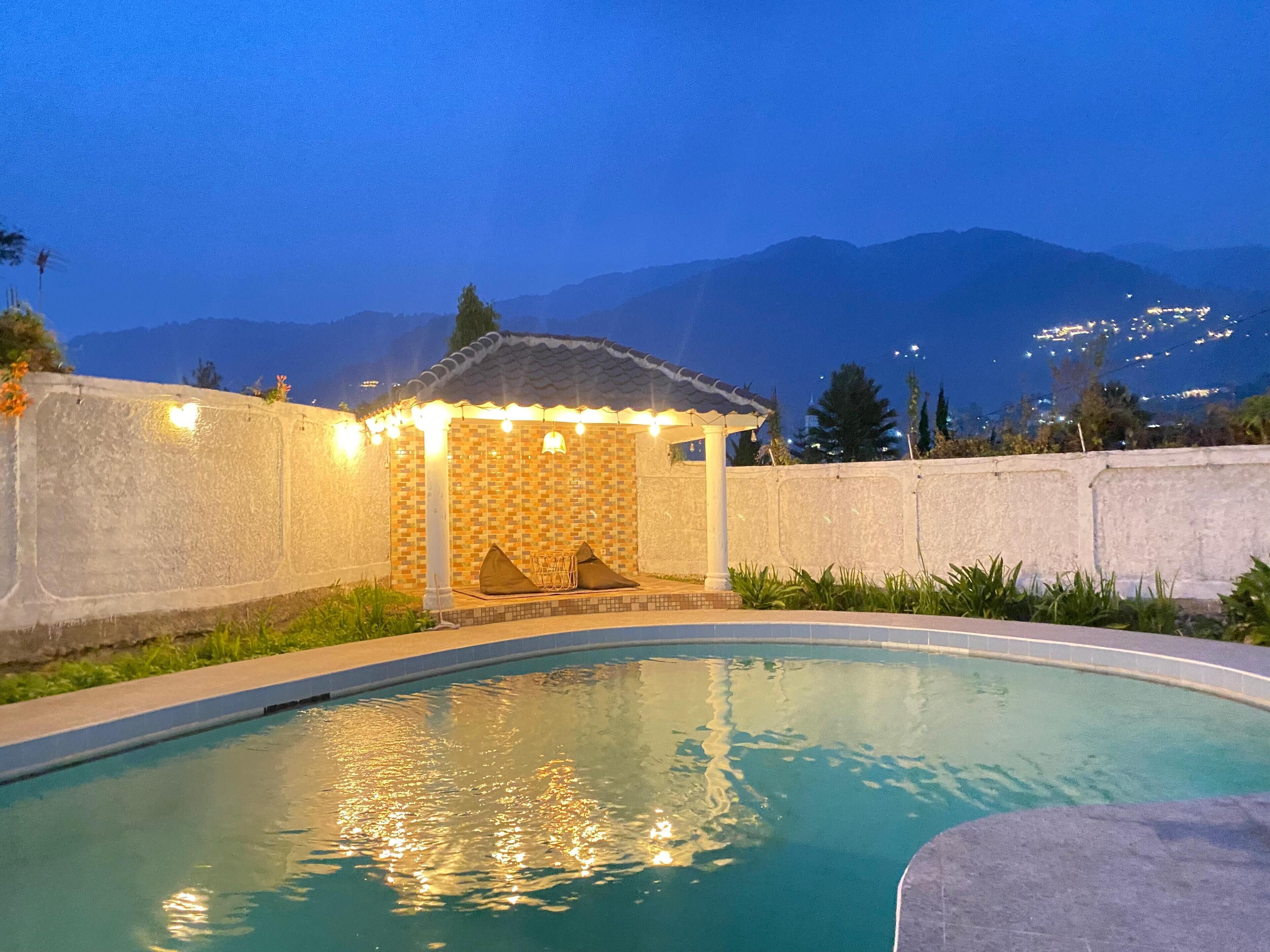 Una piscina con solárium de azulejos, zona de estar techada y vistas a montañas al fondo.
