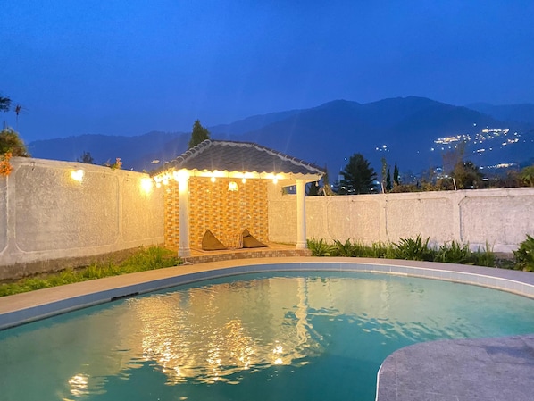 A swimming pool with a tiled deck, a covered seating area, and a view of mountains in the background.