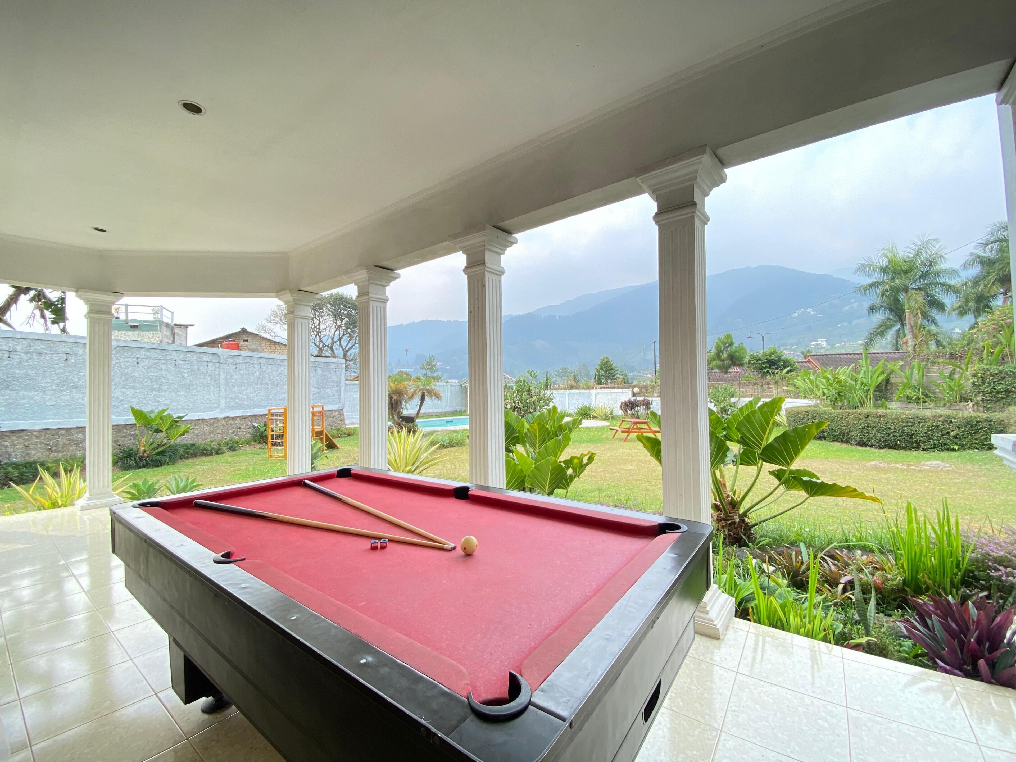 A pool table in a covered area with a view of a landscaped garden and mountains in the background.