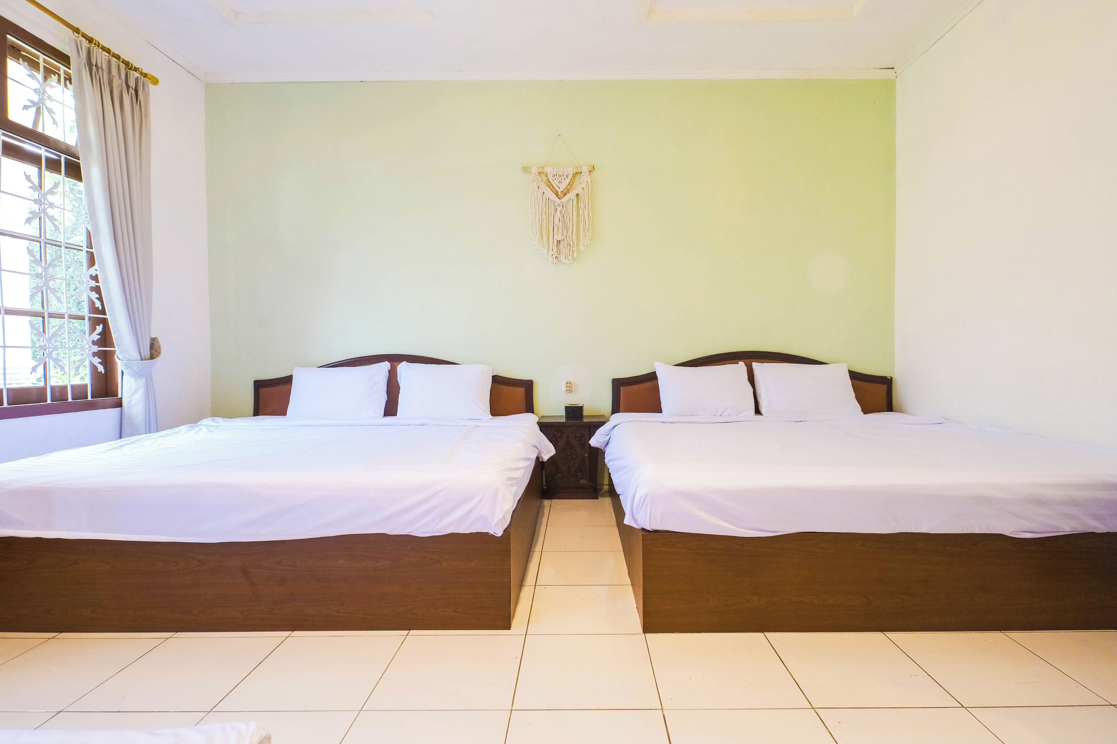 Ein Hotelzimmer mit zwei Betten, einem fensterbelaubten Fenster, einer wandhängenden Makramee-Dekoration und einem gefliesten Boden.