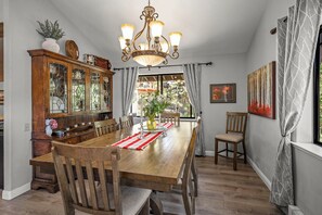 Une salle à manger avec une table en bois, des chaises, un lustre et une vue sur l’extérieur.