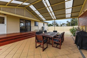 Un patio couvert avec une table et des chaises en bois, un barbecue et une porte-fenêtre coulissante donnant sur un espace intérieur.
