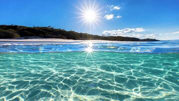 A beach with clear turquoise water, a white sandy shore, and a forested hill in the background.