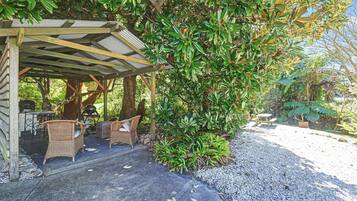 A covered outdoor seating area with wicker furniture, surrounded by lush greenery and a gravel path.