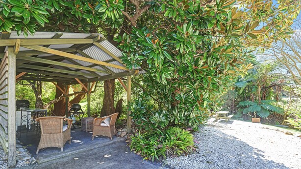 A covered outdoor seating area with wicker furniture, surrounded by lush greenery and a gravel path.