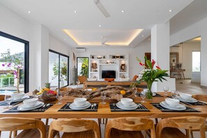 A modern dining area with a long wooden table set for a meal, a television in the background, and a view of the outdoors through large windows. - Villa Victorine (Canggu)