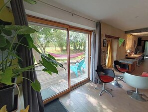 A room with a large sliding glass door leading to an outdoor area with a hammock and chairs. There are two chairs with red cushions and a modern chair with a metallic base. - Spacious house with a spa and movie theater—for families, groups, or events (Longaulnay)