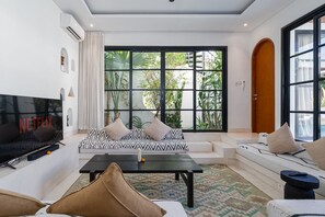 A modern living room with a flat-screen TV, a sofa set, a coffee table, and a large window with a view of greenery. - Villa Casa Libra (Pererenan)