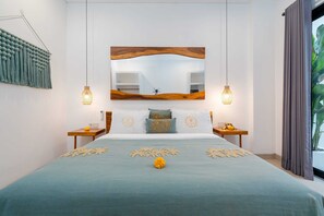 A bedroom with a bed, two bedside tables, a nightstand, a wall-mounted mirror, and a hanging light fixture. - Villa Casa Diamanti (Tumbak Bayuh)