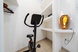 A corner of a room with a stationary bike, a wooden sculpture, and a shelf with a small item. - Villa Casa Diamanti (Tumbak Bayuh)