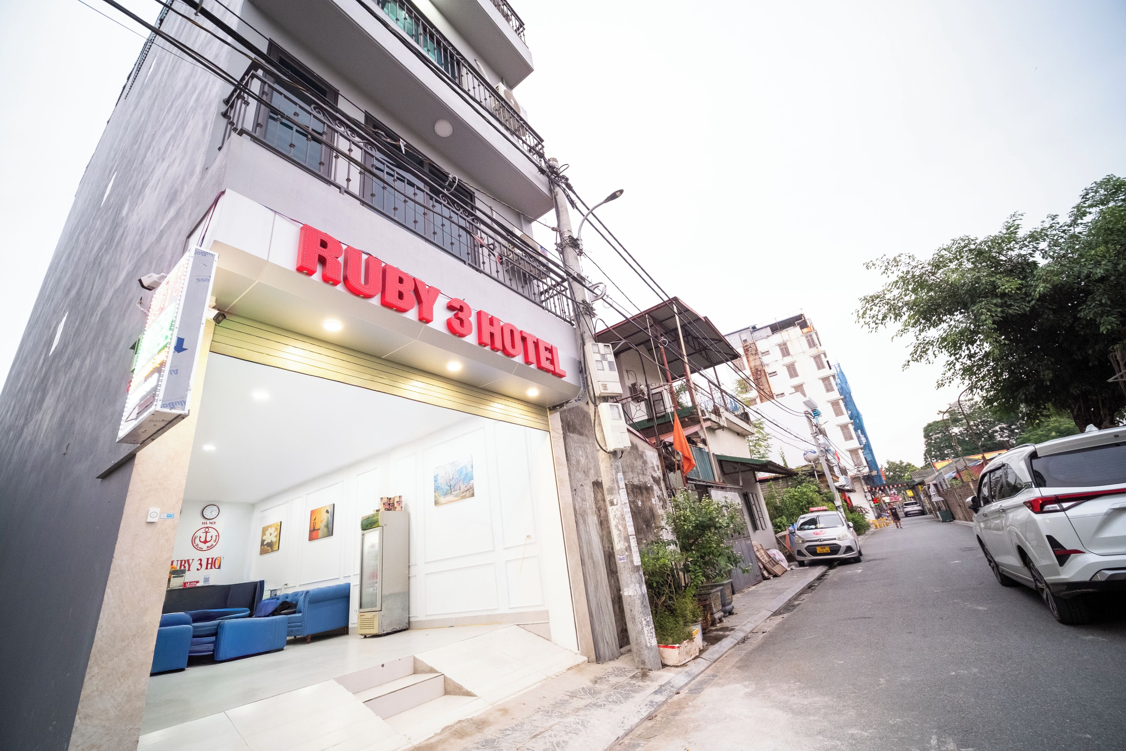 Un hotel con un llamativo letrero que dice 'Ruby 3 Hotel', una entrada visible y una vista de la calle con coches estacionados.