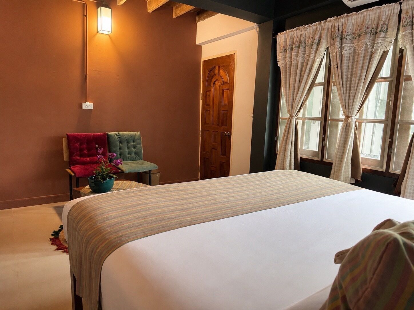 A hotel room with a bed, two chairs, a potted plant, a wooden door, and a window with curtains.