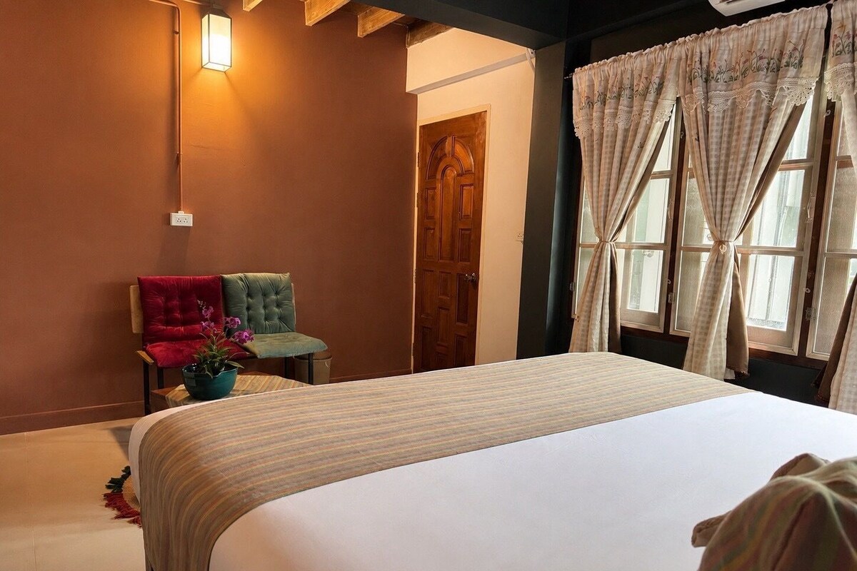 A hotel room with a bed, two chairs, a potted plant, a wooden door, and a window with curtains.