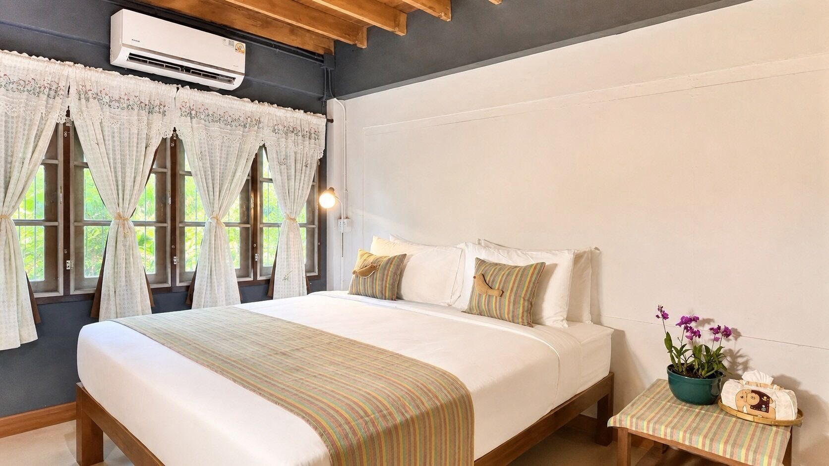 A bedroom with a bed, curtains, a bedside table with a plant, and an air conditioning unit.
