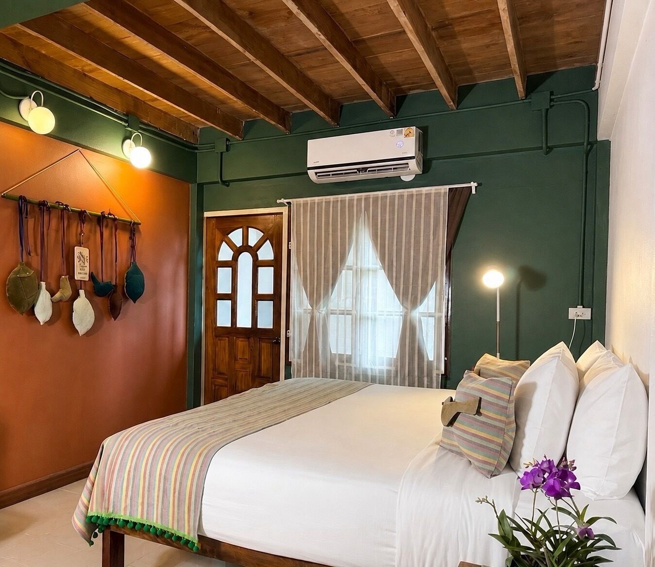 A bedroom with a bed, a window with curtains, a wall-mounted air conditioner, and a wall with hanging items.