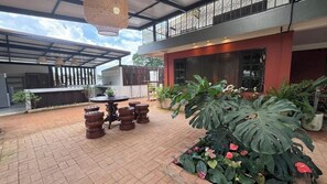 An outdoor patio area with a dining table and chairs, surrounded by potted plants and a large leafy plant.