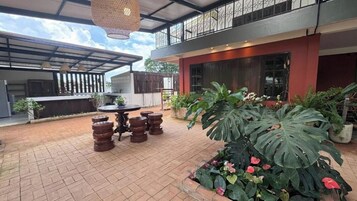 Área de terraço ao ar livre com mesa e cadeiras, rodeada por vasos de plantas e uma grande folhagem.