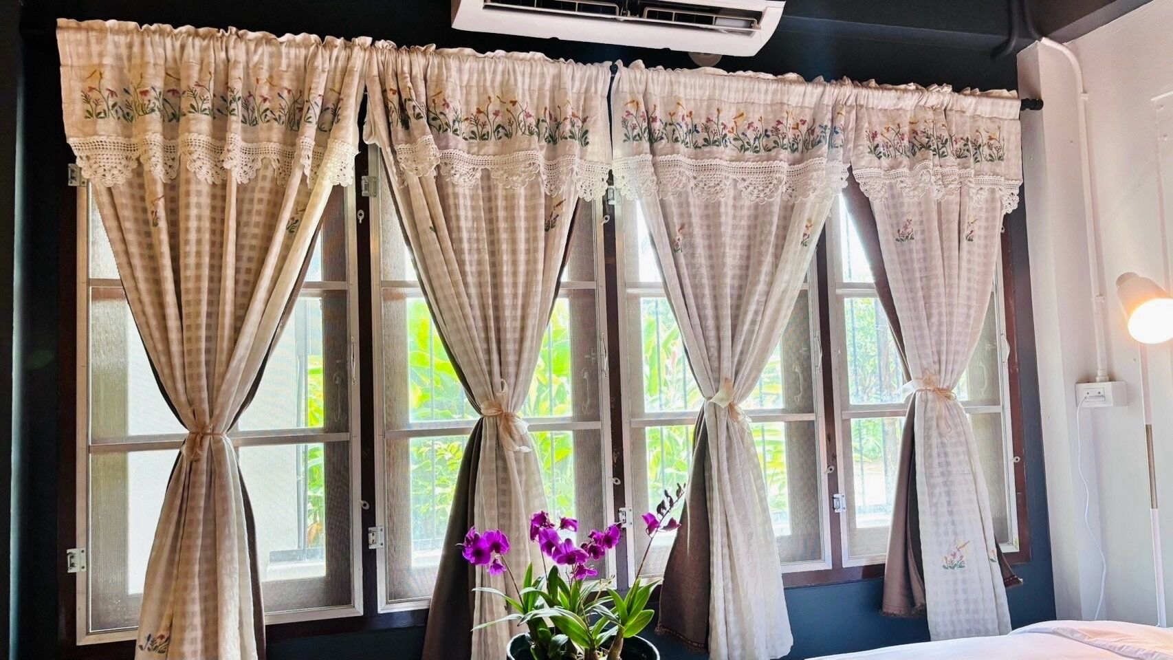 Ein Zimmer mit großem Fenster, Vorhängen mit Blumenmuster und einem Blumentopf mit violetten Blüten.