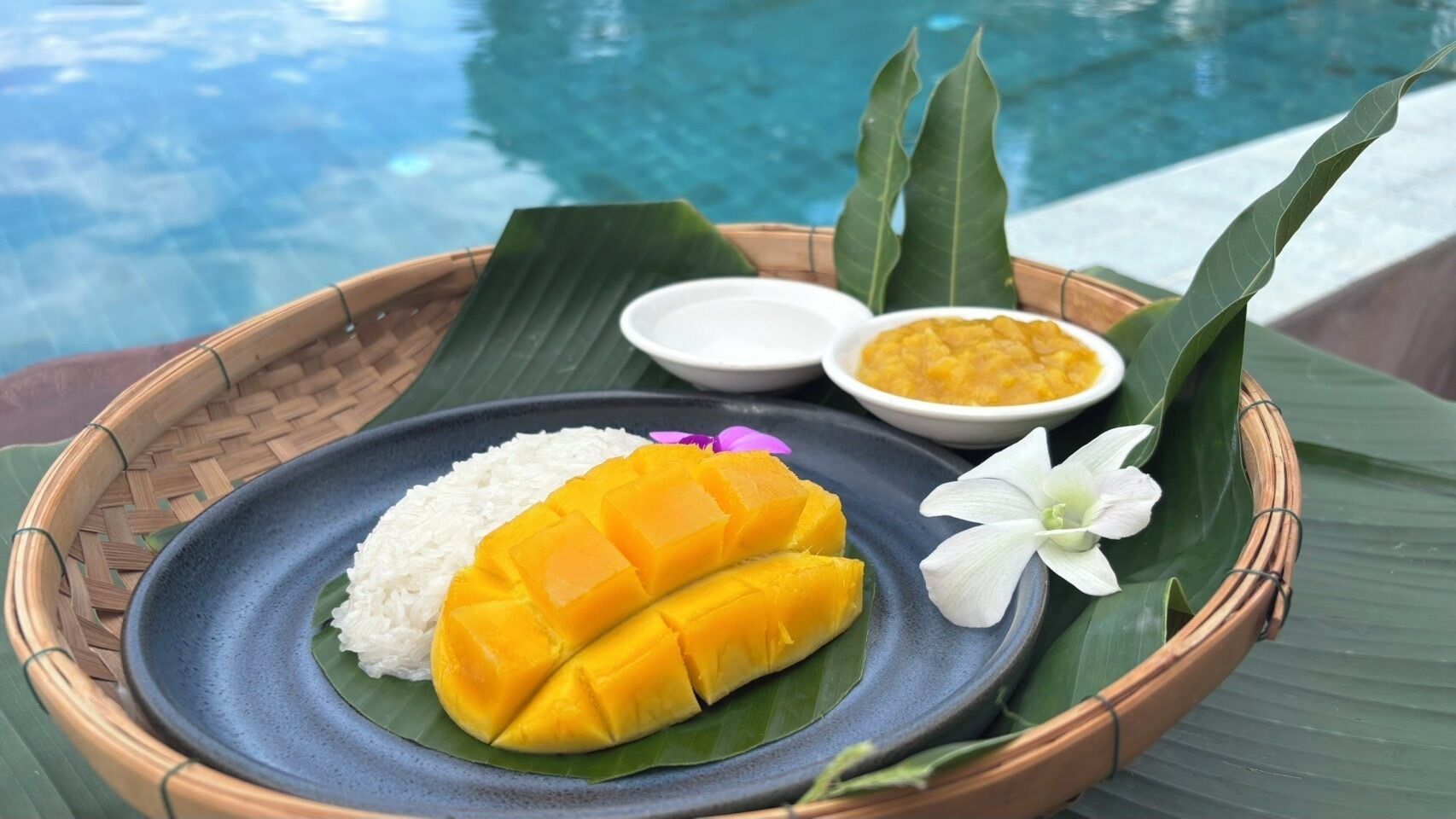 Comida que incluye arroz, tajadas de mango y un cuenco pequeño de guiso amarillo, servida en un plato azul con hoja de plátano, al lado de una piscina.