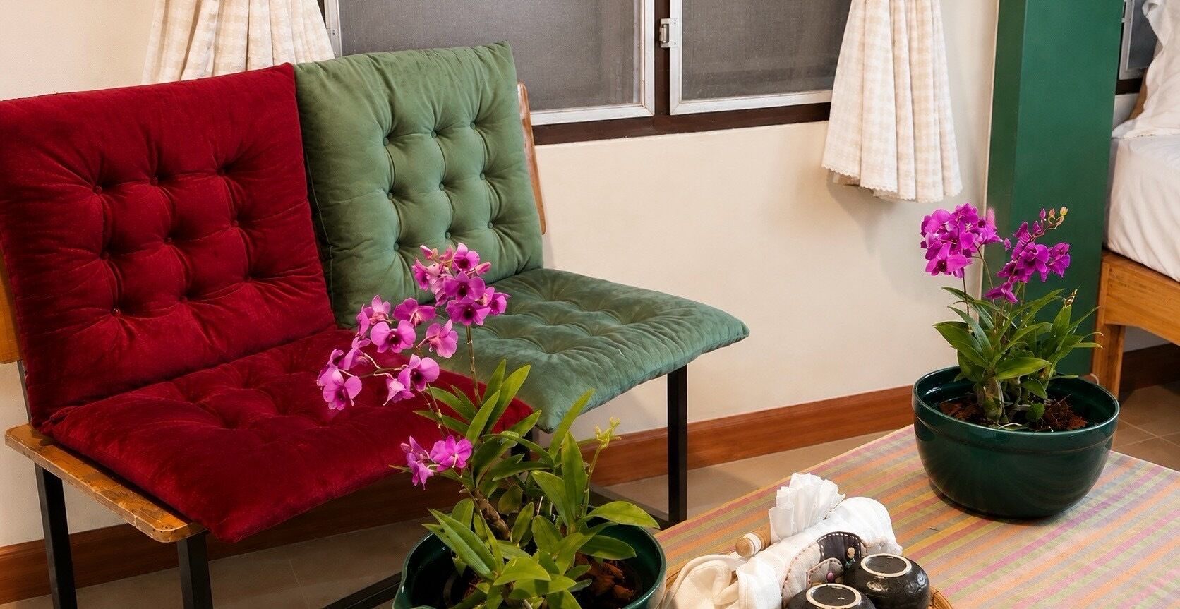 Una habitación con un sillón verde con capitoné, un sillón rojo acolchado y una mesita con una planta en maceta y una toalla blanca enrollada.