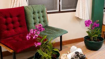 Una habitación con un sillón verde con capitoné, un sillón rojo acolchado y una mesita con una planta en maceta y una toalla blanca enrollada.