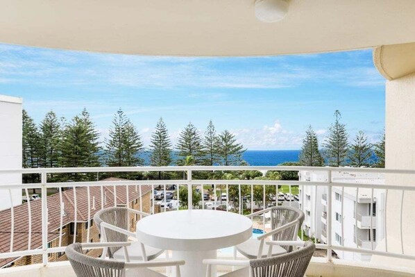 A balcony with a white table and chairs, overlooking a residential area with trees and a view of the sea. - Ocean View Coastal Haven in Burleigh Heads (Gold Coast)