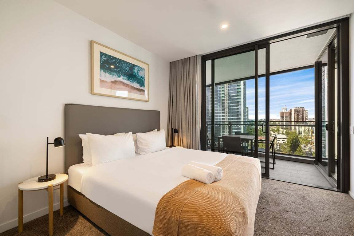 Een hotelkamer met een groot bed, een nachtkastje, een inlijsting foto aan de muur en een balkon met uitzicht over de stad.