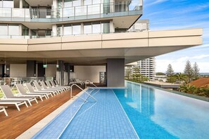 A rooftop infinity pool with lounge chairs, a modern building with balconies, and a clear sky. - The Sea-gnature Stay | Coast & City (Gold Coast)