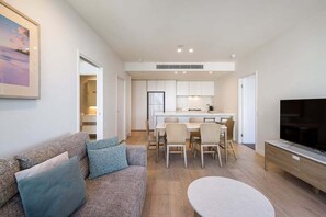 A modern living room with a sofa, a dining table, and chairs. There is a television on a stand and a kitchen area in the background. - The Sea-gnature Stay | Coast & City (Gold Coast)