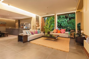 A modern living room with a sofa, coffee table, and a view of lush greenery outside. - Luxury 5-Bed Beachfront Mirage Villa with Pool (Port Douglas)